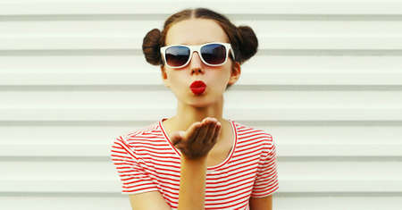 Portrait of stylish young woman blowing her lips sending sweet air kiss with red lipstick wearing a striped t-shirt with cool hairstyle on white backgroundの写真素材