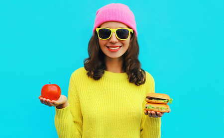 Portrait of stylish happy smiling young woman showing a burger fast food and apple on blue colorful...