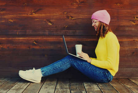 Portrait of stylish happy modern young woman drinking coffee working with laptop wearing a colorful clothes sitting in the city parkの写真素材