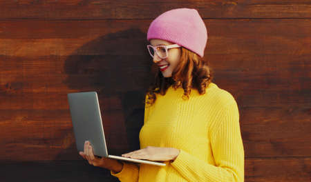 Portrait of stylish modern young woman working with laptop wearing a colorful clothes on wooden wall...