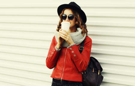 Portrait of beautiful young woman drinking coffee wearing a red jacket, black round hat on white...