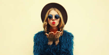 Fashionable portrait of stylish young woman, female model blowing her lips with red lipstick sending sweet air kiss wearing a blue faux fur, round hat on backgroundの写真素材