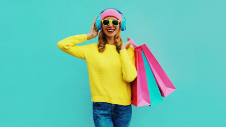 Colorful portrait of stylish smiling young woman listening to music in headphones with shopping bags posing wearing a yellow knitted sweater, pink hat on blue backgroundの写真素材