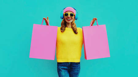 Colorful portrait of stylish smiling young woman listening to music in headphones with shopping bags posing wearing a yellow knitted sweater, pink hat on blue backgroundの写真素材