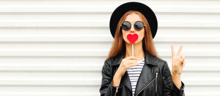 Fashionable portrait of stylish young woman with red heart shaped lollipop blowing her lips sending...