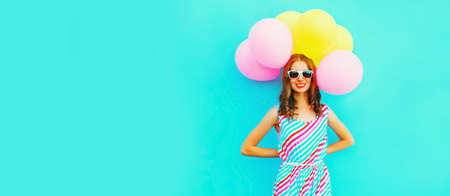 Portrait of beautiful happy smiling young woman with bunch of balloons wearing a colorful dress on blue backgroundの写真素材