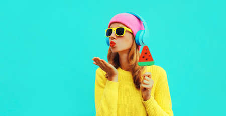 Summer fresh colorful portrait of happy young woman in headphones listening to music with fruit juicy lollipop or ice cream shaped slice of watermelon on blue backgroundの写真素材