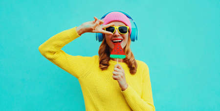 Summer fresh colorful portrait of happy laughing young woman in headphones listening to music with fruit juicy lollipop or ice cream shaped slice of watermelon on blue backgroundの写真素材