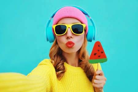 Summer fresh colorful portrait of young woman taking selfie in headphones listening to music with fruit juicy lollipop or ice cream shaped slice of watermelon on blue backgroundの写真素材