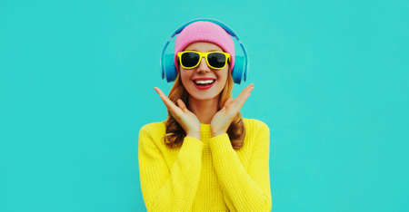 Portrait of happy surprised smiling young woman model with headphones listening to music wearing colorful yellow sweater on blue backgroundの写真素材