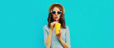 Portrait of stylish young woman drinking a fresh juice wearing a summer straw hat and striped t-shirt on blue backgroundの写真素材