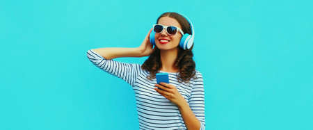 Portrait of smiling young woman in headphones listening to music with smartphone on blue backgroundの写真素材
