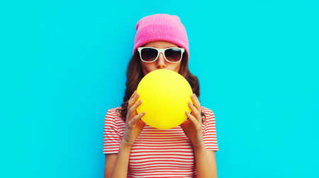 Fashionable portrait of stylish cool young woman inflating chewing gum wearing pink hat on blue...