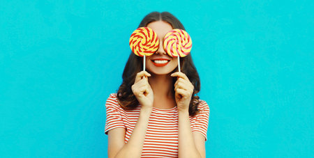 Portrait of happy smiling young woman covering her eyes with lollipop on blue background