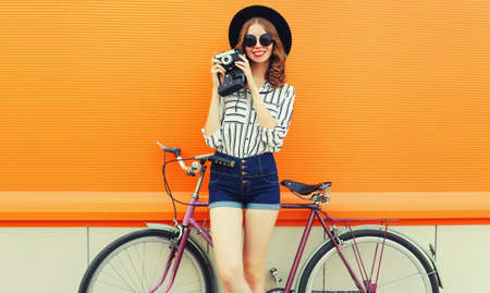 Summer colorful portrait of happy smiling young woman photographer taking a picture by film camera with bicycle in the city on orange backgroundの写真素材