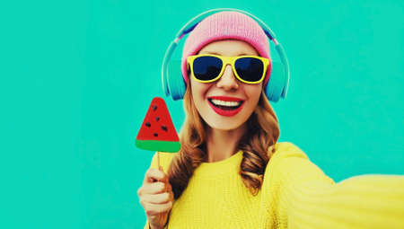 Summer fresh colorful portrait of laughing woman model taking selfie in headphones listening to music with fruit juicy lollipop or ice cream shaped slice of watermelon on blue backgroundの写真素材