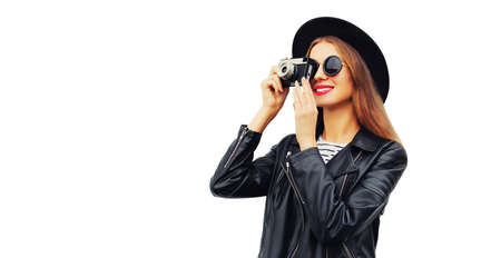 Stylish portrait of beautiful young woman photographer taking picture with film camera wearing black round hat, leather jacket isolated on white backgroundの写真素材