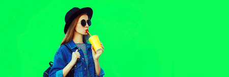 Young woman drinking juice looking away wearing black round hat, denim jacket on green background, blank copy space for advertising textの写真素材