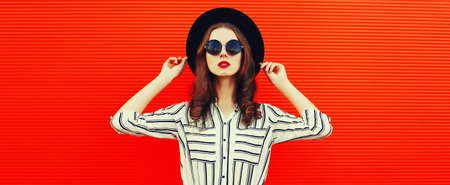 Portrait of beautiful young woman wearing white striped shirt, black round hat on red backgroundの写真素材