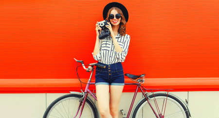 Summer image of happy smiling young woman photographer taking a picture by film camera with bicycle in the city on red backgroundの写真素材