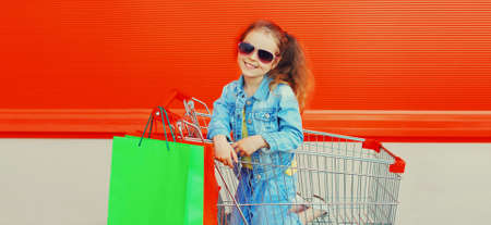 Portrait of happy smiling little girl child with trolley cart and shopping bags wearing denim clothes on city streetの写真素材
