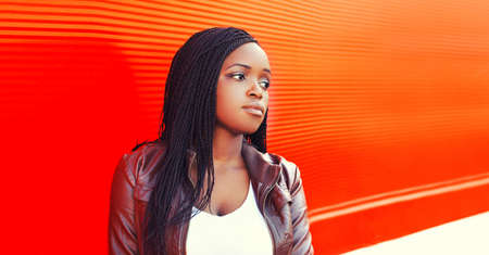 Portrait of stylish african young woman with dreadlocks looking away wearing brown jacket on red...
