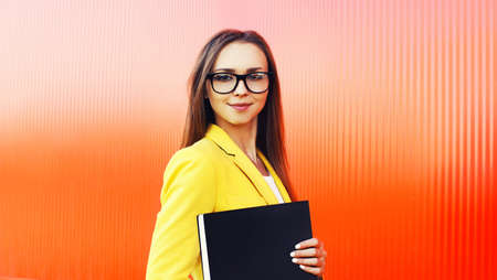 Portrait of young woman with folder wearing eyeglasses, yellow business suit on vivid background