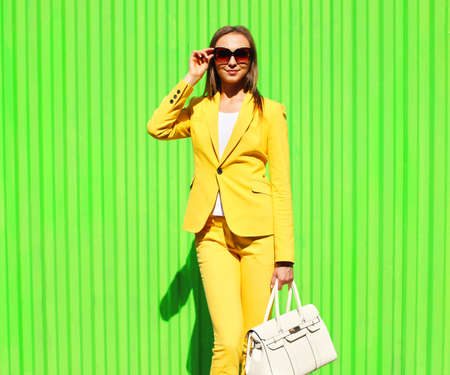 Beautiful young woman wearing yellow suit with handbag posing on vivid green background, street fashionの写真素材