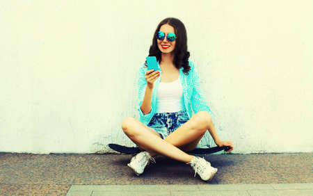 Modern young woman with smartphone sitting on skateboard on city street over white background
