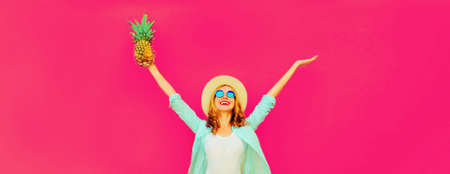 Summer vacation, happy laughing woman raising her hands up with pineapple having fun wearing straw hat on pink backgroundの写真素材