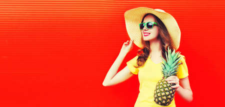 Summer portrait of happy smiling woman with pineapple looking away wearing summer straw hat on red background, blank copy space for advertising textの写真素材