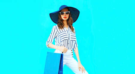 Portrait of beautiful young woman with shopping bags wearing white striped shirt, summer black round straw hat on blue backgroundの写真素材
