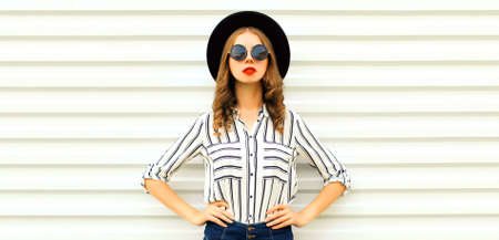 Portrait of beautiful young woman wearing striped shirt and black round hat on white background,...