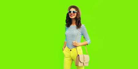 Portrait of happy smiling stylish young woman posing wearing handbag, striped t-shirt and summer straw round hat on green background, blank copy space for advertising textの写真素材