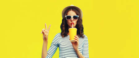 Portrait of stylish young woman drinking fresh juice wearing summer straw hat, sunglasses and striped t-shirt on yellow backgroundの写真素材