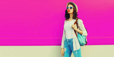Beautiful young woman posing wearing summer straw hat, backpack on city street on pink background,...