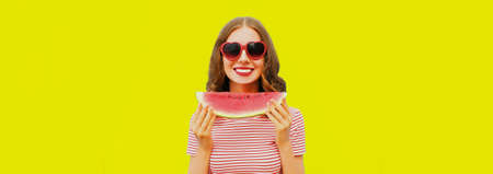 Portrait of happy smiling young woman with slice of watermelon wearing red heart shaped sunglasses on yellow background, blank copy space for advertising textの写真素材