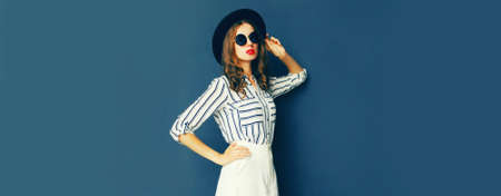 Portrait of beautiful young woman wearing white striped shirt, black round hat on dark blue backgroundの写真素材