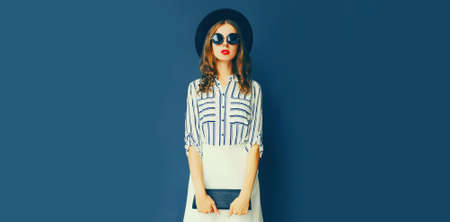 Full length beautiful young woman wearing white striped shirt, black round hat, handbag clutch on dark blue backgroundの写真素材