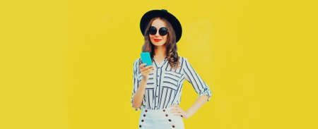 Portrait of stylish woman with smartphone wearing black round hat, white striped shirt on yellow backgroundの写真素材
