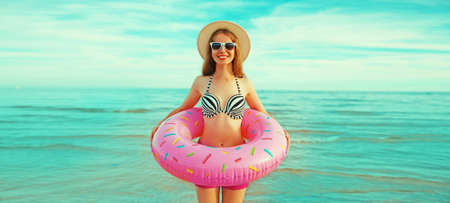 Portrait of happy smiling young woman with inflatable ring wearing straw hat on beach on sea backgroundの写真素材