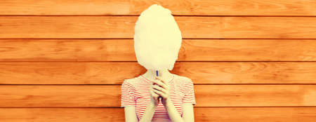 Portrait of woman having fun covering her face with cotton candy in amusement park on wooden wall backgroundの写真素材