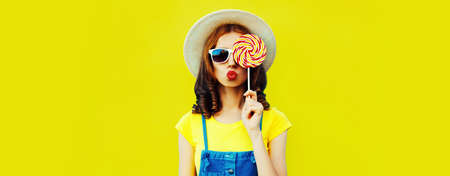 Summer portrait of stylish young woman with lollipop blowing her lips sending sweet air kiss wearing straw hat on yellow backgroundの写真素材