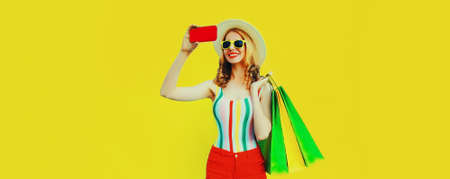 Portrait of happy smiling young woman with shopping bags taking selfie with smartphone wearing summer straw hat on yellow backgroundの写真素材
