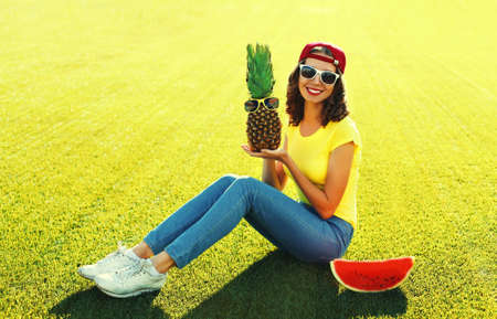 Summer portrait of happy smiling young woman with pineapple and fresh juicy slice of watermelon on the grass in the summer parkの写真素材