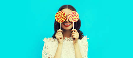 Portrait of cheerful smiling young woman covering her eyes with lollipop on blue backgroundの写真素材