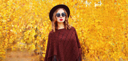 Portrait of stylish young woman wearing black round hat and knitted brown poncho in the park on...