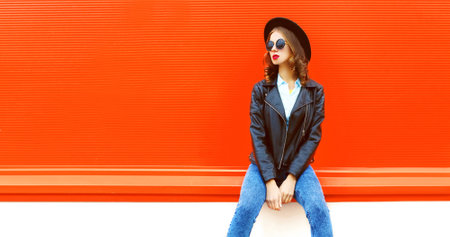 Portrait of stylish woman model posing wearing black rock style leather jacket, round hat on red backgroundの写真素材