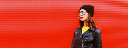 Portrait of stylish woman model posing wearing black rock style leather jacket, hat, eyeglasses on red backgroundの写真素材