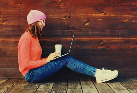Portrait of stylish happy modern young woman drinking coffee working with laptop wearing a colorful clothes sitting in the city parkの写真素材